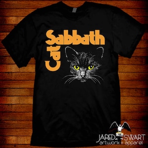 May include: Black t-shirt featuring a graphic of a black cat with yellow eyes. The text "Sabbath Cat" is printed in orange above the cat's face. The shirt is a classic crew neck style.