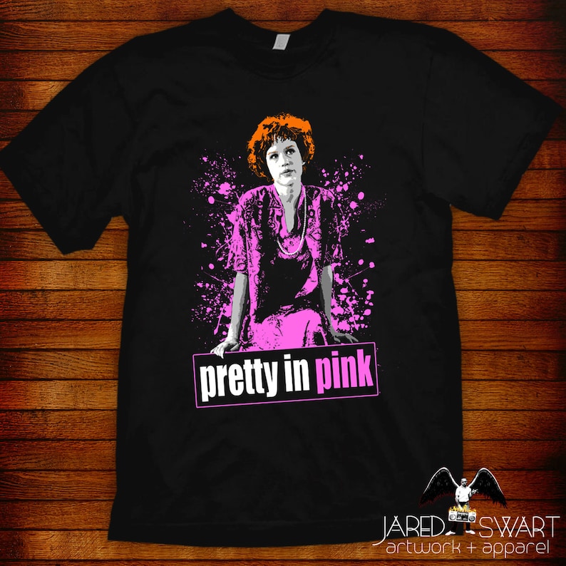 May include: Black t-shirt with a pink and white graphic of a woman with red hair. The graphic has a pink splatter effect and the text "pretty in pink" in white.