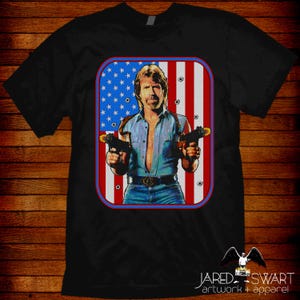 May include: Black t-shirt featuring a graphic of a man holding two guns in front of an American flag design. The graphic is framed in a red border. The shirt has the text "JARED SWART artwork + apparel" at the bottom.