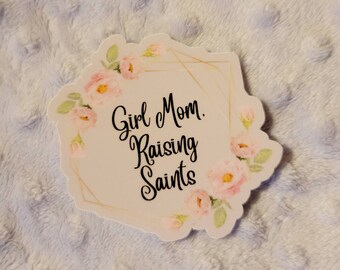 May include: A white sticker with a gold geometric frame and pink floral accents. The text "Girl Mom. Raising Saints" is written in black cursive font.