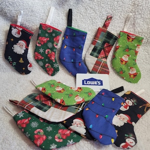 May include: A collection of small, handmade Christmas stockings in various festive patterns. Designs include Santa Claus, candy canes, Christmas lights, and plaid. Each stocking has a loop for hanging and is made of fabric in colors like red, green, and blue.