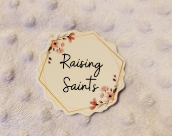 May include: A white hexagonal sticker with a gold outline and floral accents. The sticker says "Raising Saints" in black cursive font.