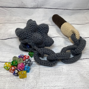 Könnte beinhalten: Ein grauer Häkel-Würfelbeutel mit einer Kette und einem braunen und beigefarbenen Häkelgriff. Der Beutel ist mit bunten Würfeln gefüllt.