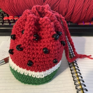 May include: A red and green crocheted watermelon-shaped drawstring bag with black beads. The bag has a white stripe between the red and green sections.