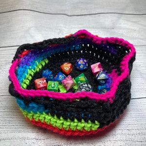 May include: A crocheted dice bag with a black, green, blue, red, and pink color scheme. The bag is filled with colorful dice.