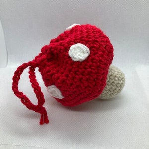 May include: A red crocheted mushroom with white polka dots and a beige stem. The mushroom has a drawstring.