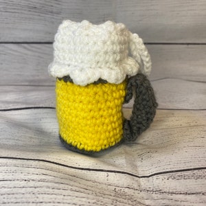 May include: A crocheted beer mug with a white foam top, yellow beer, and a gray handle.