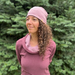 May include: A woman with curly brown hair is wearing a mauve cowl neck top and a mauve beanie with a small brown tag. She is standing in front of a green leafy background.