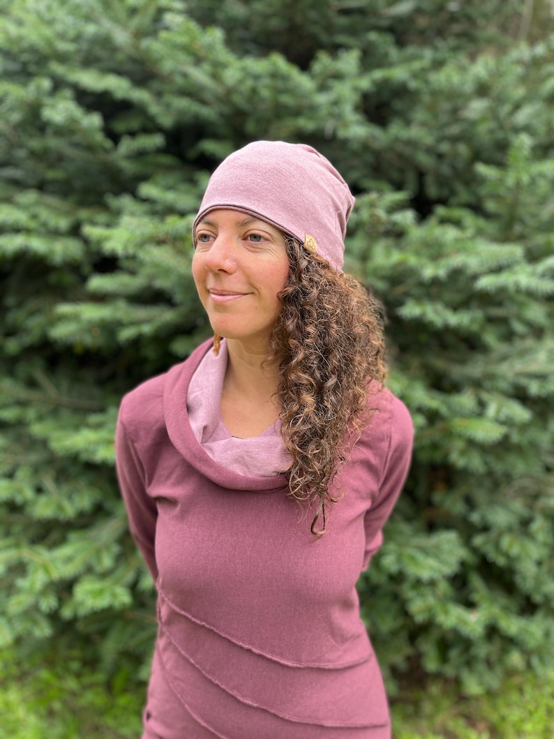 May include: A woman with curly brown hair is wearing a mauve cowl neck top and a mauve beanie with a small brown tag. She is standing in front of a green leafy background.