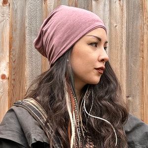 May include: A woman with long brown hair is wearing a mauve beanie hat. She is wearing a black jacket with a brown and white patterned lining. She has feather hair extensions in her hair.
