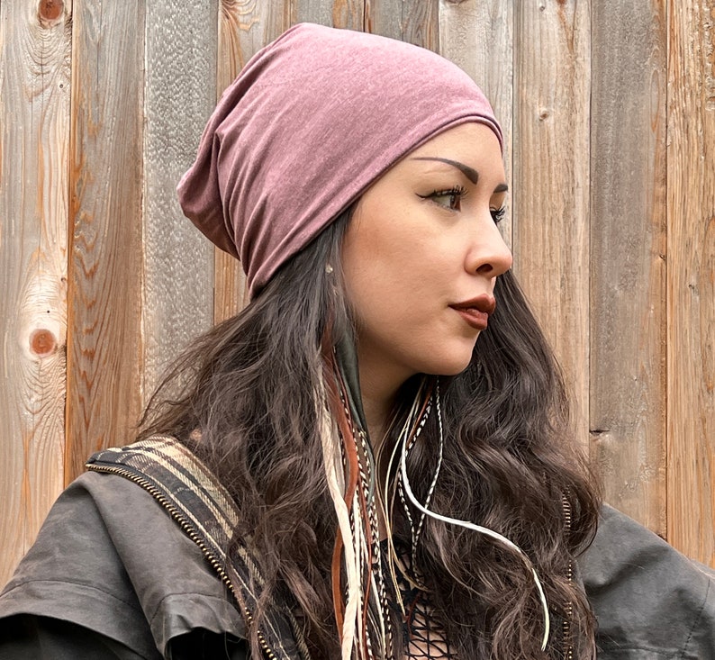 May include: A woman with long brown hair is wearing a mauve beanie hat. She is wearing a black jacket with a brown and white patterned lining. She has feather hair extensions in her hair.