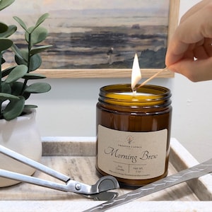 Op de afbeelding: Een brandende bruine glazen potkaars met het label "Morning Brew". Een zilveren lonttrimmer en een decoratieve metalen plaat liggen op een marmeren dienblad. Links staat een kleine potplant.