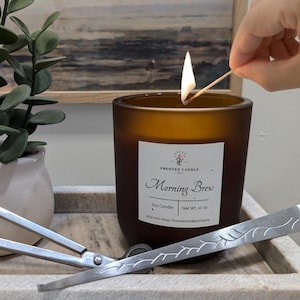 Op de afbeelding: Een bruine, gematteerde glazen kaars met een wit etiket waarop "Frosted Candle Co." en "Morning Brew Soy Candle Net Wt 10 oz" staat. De kaars wordt aangestoken met een lucifer. Er liggen twee zilveren kaarsendoekjesnijders op een wit marmeren dienblad naast de kaars.
