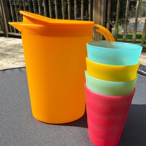 May include: An orange plastic pitcher with a lid and handle stands next to a stack of four colorful cups. The cups are stacked in shades of red, green, yellow, and blue. The pitcher and cups are made of plastic.