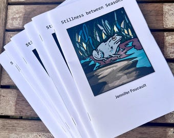 Könnte beinhalten: Ein Stapel weißer Broschüren mit dem Titel "Stillness between Seasons" und dem Namen der Autorin, Jennifer Foucault. Das Cover zeigt einen Frosch in einem Teich. Die Broschüren liegen auf einer Holzoberfläche.
