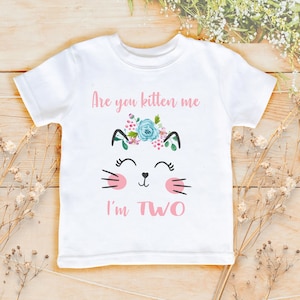 May include: White t-shirt with a pink cat graphic and the text "Are you kitten me I'm TWO". The cat has a floral crown and pink cheeks.