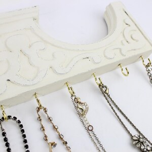 May include: White wooden wall-mounted jewelry organizer with gold hooks and various necklaces and bracelets hanging from them.
