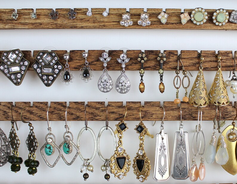 May include: A wooden earring holder with three rows of earrings. The earrings are a variety of styles, including silver, gold, and turquoise. Some earrings feature crystals, others have beads, and some have a more rustic look.