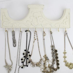 May include: White wooden wall mounted jewelry organizer with ornate detailing.  The organizer has hooks for hanging necklaces and earrings.  There are six necklaces hanging from the hooks, including a black beaded necklace, a silver chain necklace with a flower pendant, and a gold chain necklace with a pearl pendant.
