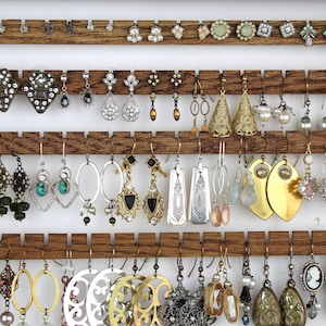 May include: A wooden jewelry organizer rack with four horizontal slats featuring notched edges to hold various pairs of earrings. The collection includes silver, gold, and bronze-toned dangles, hoops, studs, and cameo-style earrings.