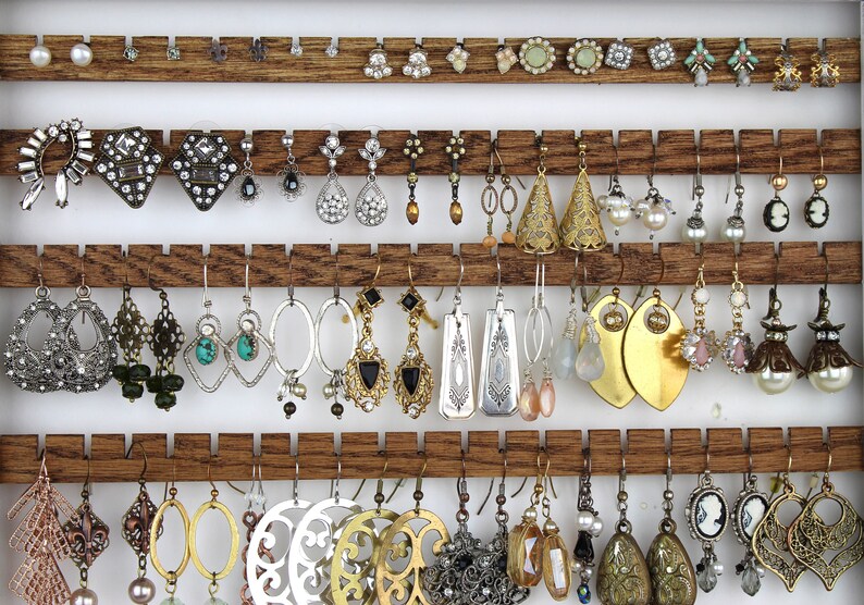 May include: A wooden jewelry organizer rack with four horizontal slats featuring notched edges to hold various pairs of earrings. The collection includes silver, gold, and bronze-toned dangles, hoops, studs, and cameo-style earrings.