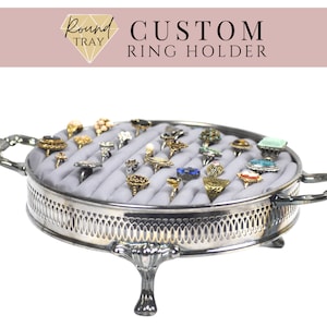May include: Silver ring holder tray with a gray velvet lining and a variety of rings with different colored stones. The tray has a decorative border and three legs.