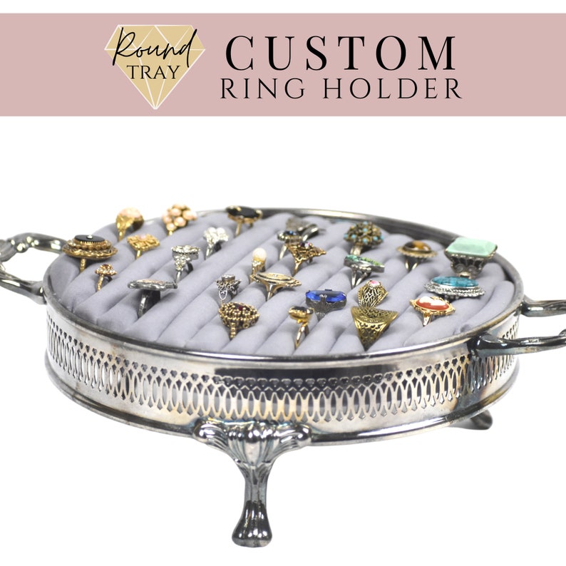 May include: Silver ring holder tray with a gray velvet lining and a variety of rings with different colored stones. The tray has a decorative border and three legs.