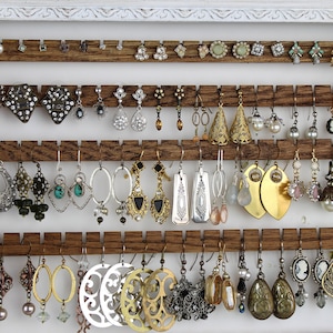 May include: A white wooden frame with a brown wooden backing holds a collection of earrings. The earrings are arranged in rows on hooks. The earrings are made of various materials, including metal, glass, and beads.