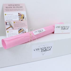 May include: A white rectangular box topped with a pink paper roll tied with string, featuring a Creatively Cluttered brand label. An instructional card titled Attaching Slats to Frame with numbered steps and photos is included alongside the box.