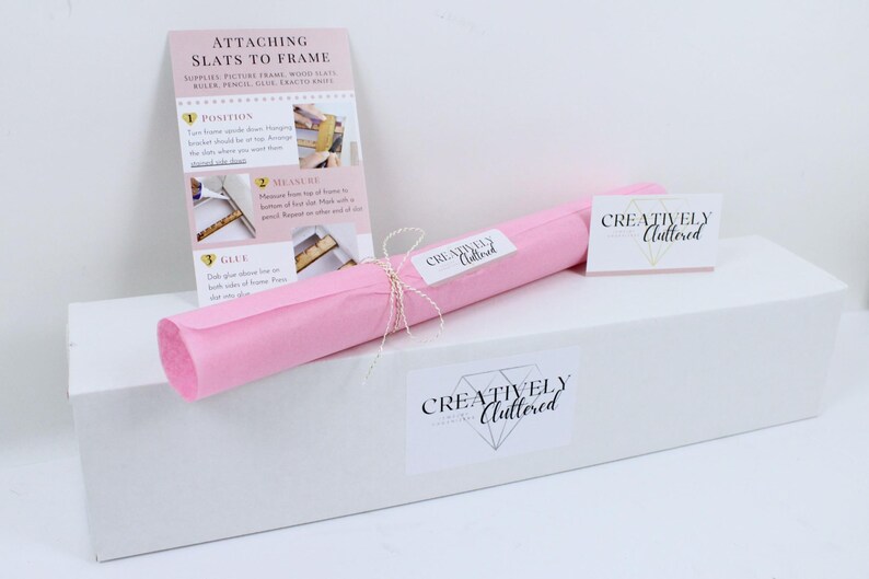 May include: A white rectangular box topped with a pink paper roll tied with string, featuring a Creatively Cluttered brand label. An instructional card titled Attaching Slats to Frame with numbered steps and photos is included alongside the box.