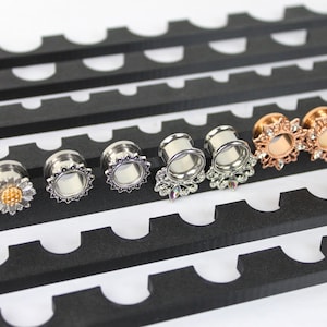 May include: An assortment of ear gauges in various designs, including silver, gold, and black. Some gauges feature floral and sunburst patterns, while others have a simple, sleek design. The gauges are displayed on a black, slotted display.