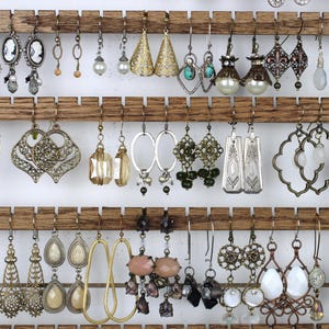 May include: A selection of earrings in various styles and colours, including gold, silver, and bronze. The earrings feature pearls, gemstones, and detailed metalwork. The collection is displayed on a wooden rack, showcasing a range of designs.