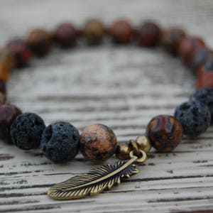 May include: A beaded bracelet featuring a mix of brown, orange, and black stone beads. The bracelet includes a gold-colored feather charm. The beads vary in size and texture, creating a natural, earthy aesthetic.