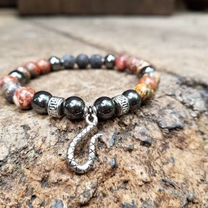 May include: A beaded bracelet featuring a mix of colorful jasper and dark gray hematite beads. The bracelet has a silver-toned snake charm dangling from the center. The beads are strung on an elastic cord, creating a stretchable design.