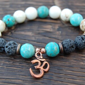 May include: A beaded bracelet featuring a copper Om charm. The bracelet includes white, turquoise, and black lava stone beads. The beads are strung together with a copper-colored spacer. The bracelet is displayed on a wooden surface.