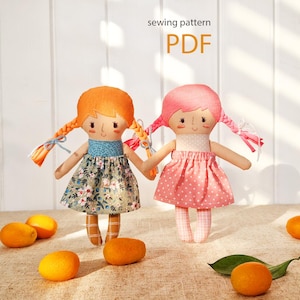 Puede incluir: Dos muñecas de tela hechas a mano con trenzas, una con cabello naranja y un vestido floral, la otra con cabello rosa y un vestido de lunares rosas. Las muñecas se toman de la mano. La imagen incluye el texto "sewing pattern PDF" y varias naranjas.