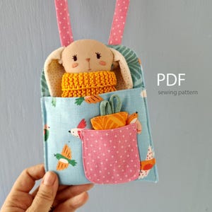 Puede incluir: Un juguete de conejito de tela hecho a mano con una bufanda amarilla tejida, metido en una bolsa azul con un bolsillo rosa. La bolsa presenta un patrón de pájaros y flores, con una correa rosa. El texto "PDF sewing pattern" es visible.