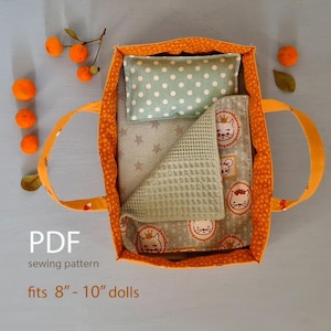 May include: A handmade doll bed with an orange and white patterned exterior, featuring a light green polka dot pillow, a grey star-patterned blanket, and a light green waffle-textured blanket. The bed has orange handles. The text "PDF sewing pattern fits 20-25 cm dolls" is visible.