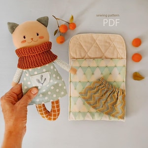 Puede incluir: Una muñeca de gato hecha a mano con una bufanda color óxido y un vestido verde claro con lunares. La muñeca está junto a una cama y unos pantalones cortos a juego, con el texto "sewing pattern PDF".