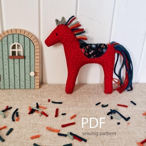 Op de afbeelding: Een handgemaakt rood stoffen paard met een bloemenzadel en een kleurrijke manen en staart. Het paard staat naast een kleine houten deur. De afbeelding bevat ook de tekst "PDF sewing pattern".