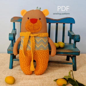 May include: A handmade brown bear plushie with a red nose, yellow ears, and a yellow and blue patterned scarf. The bear is wearing orange pants with a white and orange pattern. The image includes the text "PDF sewing pattern".