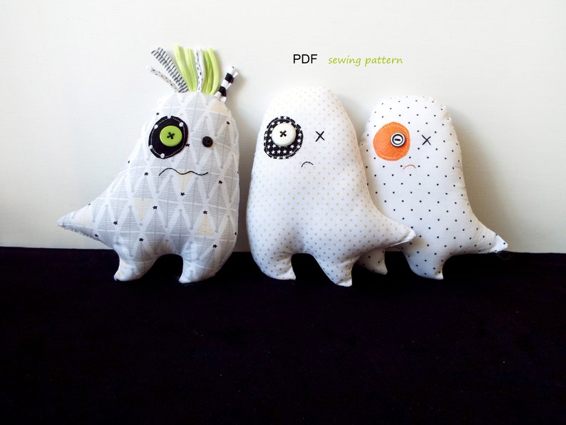 May include: Three handmade monster-shaped plush toys. The first is white with a geometric pattern, the second is white with gold polka dots, and the third is white with black polka dots. Each has button eyes and stitched details. The text "PDF sewing pattern" is visible.