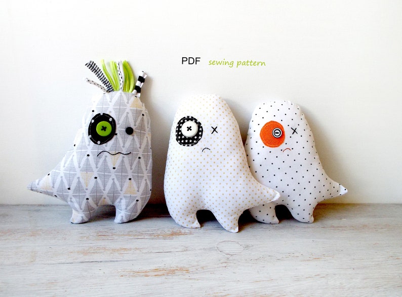 May include: Three handmade monster plushies. The first is gray and white patterned with a green button eye and black button. The second is white with gold dots and a black and white button eye. The third is white with black dots and an orange button eye. Text: PDF sewing pattern.