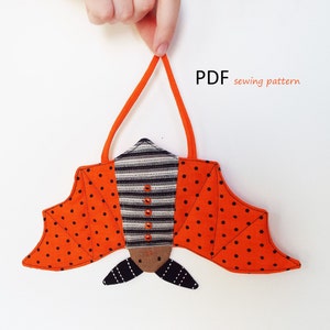 May include: A PDF sewing pattern for a Halloween bat ornament. The bat is orange with black polka dots and has a striped body. The bat is hanging from a loop of orange fabric.