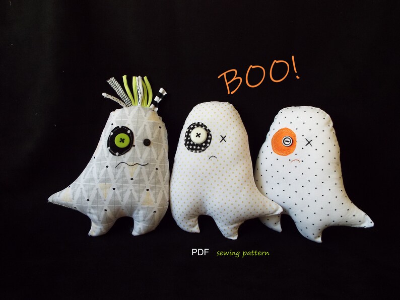 May include: Three handmade ghost plushies with button eyes and stitched mouths are displayed against a black background. One ghost has a patterned body, while the others have polka dots. The word "BOO!" is written above them.