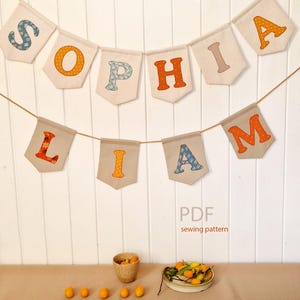 Può includere: Un banner che dice "SOPHIA" e "LIAM" con lettere colorate. Le lettere sono cucite su bandierine beige e infilate su un cordoncino marrone. Le parole sono visualizzate contro un muro bianco. L'immagine include anche una piccola ciotola di arance e un piatto di frutta.