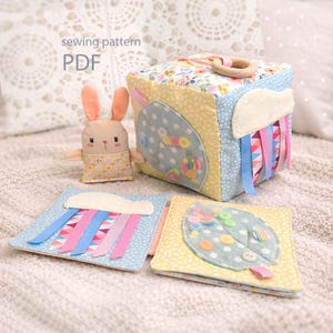 May include: A handmade fabric baby toy set, including a cube, two small books, and a bunny. The cube features a wooden ring, colourful ribbons, and leaf-shaped appliqués with buttons. The books have cloud and leaf designs. The text "sewing pattern PDF" is visible.