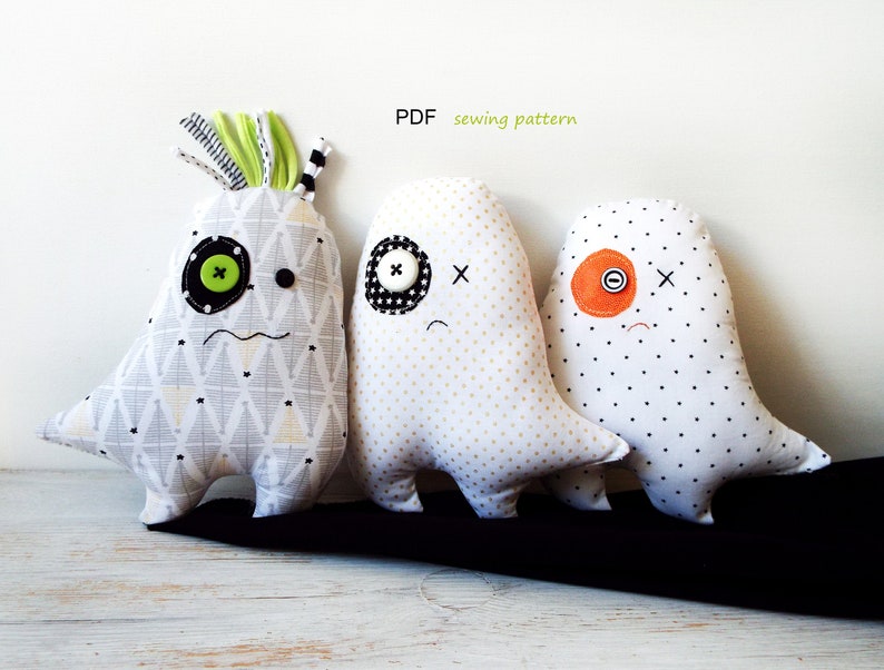 May include: Three handmade fabric monster dolls with button eyes and stitched mouths. One doll has a green button eye and a patterned fabric body. The other two dolls have black and orange button eyes and white dotted fabric bodies. The text "PDF sewing pattern" is visible.