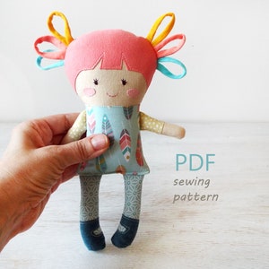 Puede incluir: Patrón de costura PDF para una muñeca hecha a mano con cabello rosa, coletas azules y amarillas, un vestido turquesa con estampado de plumas y pantalones con estampado gris y blanco.