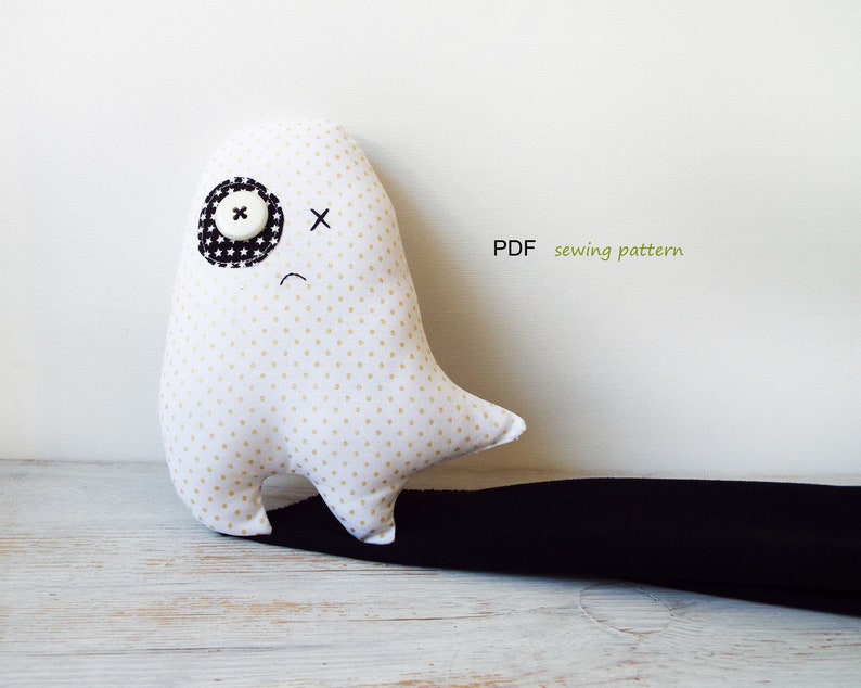 May include: A white ghost-shaped plush toy with gold polka dots, a black and white star-patterned eye patch, and a button eye. The ghost has a stitched black cross for an eye and a stitched curved mouth. The text "PDF sewing pattern" is visible.
